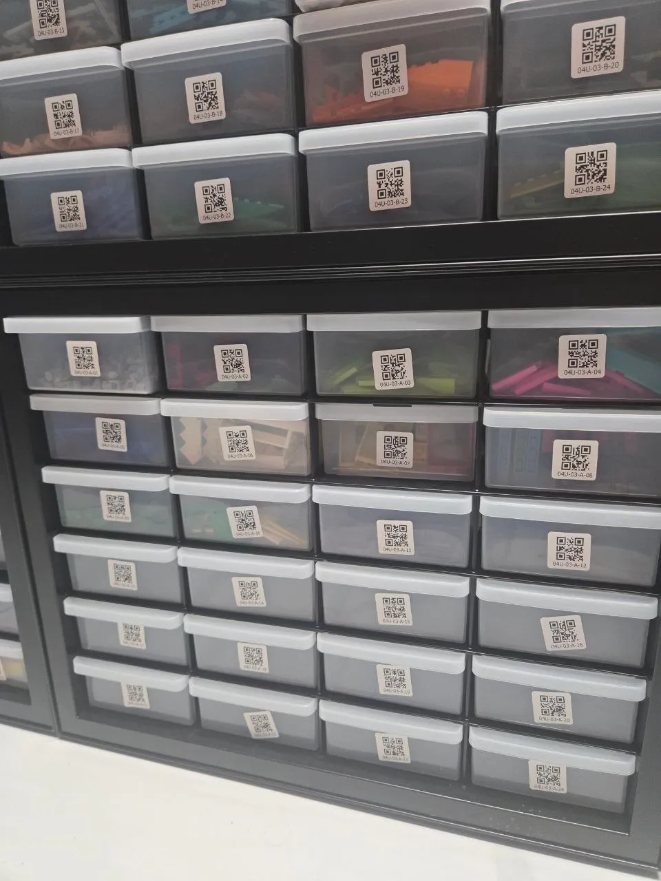Stacked black 24-drawer cabinets with QR-code location labels on clear drawers of colorful LEGO parts