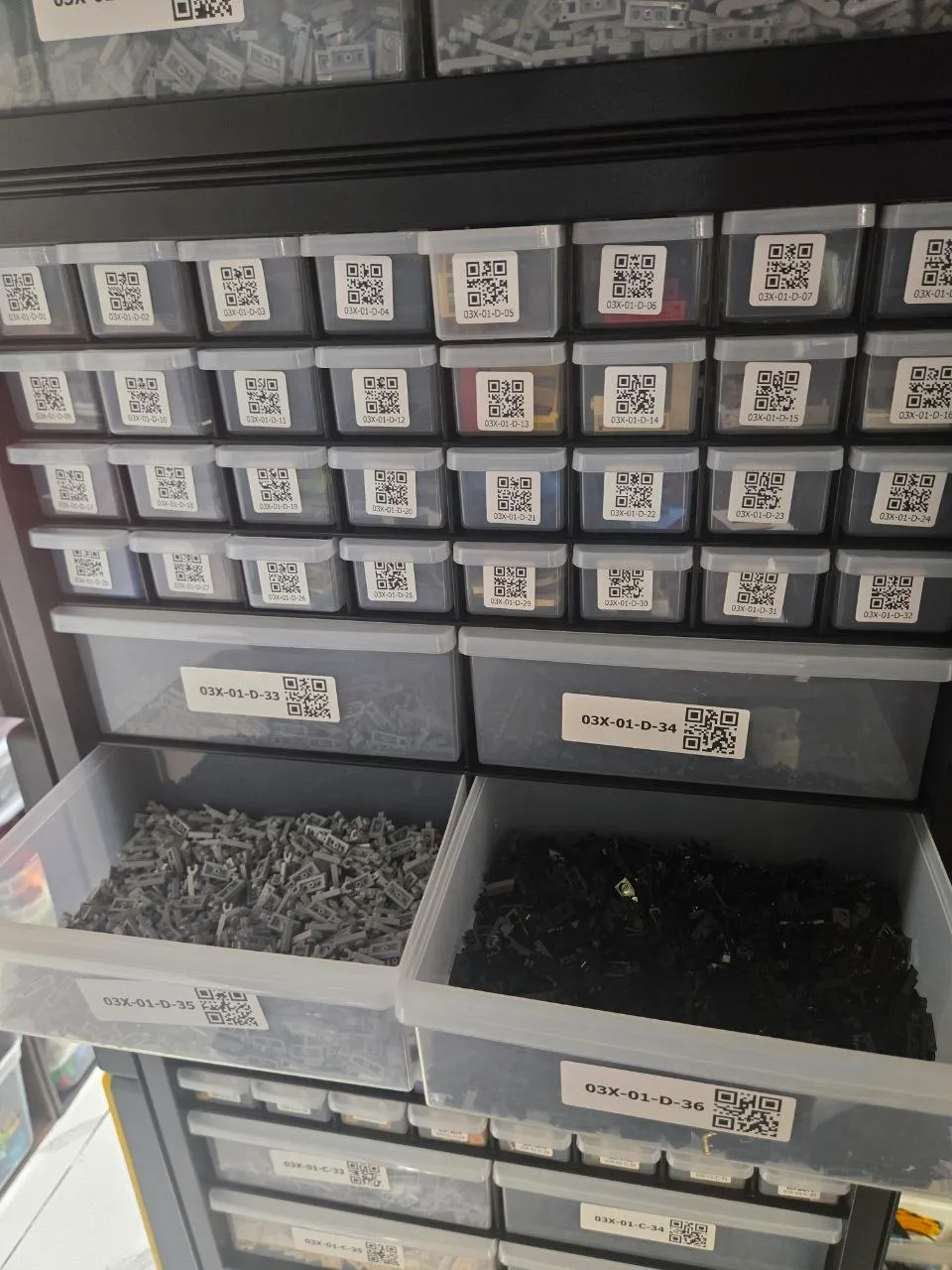 Two large drawers of dark gray and black LEGO parts pulled out of a black 36-drawer cabinet with QR labels