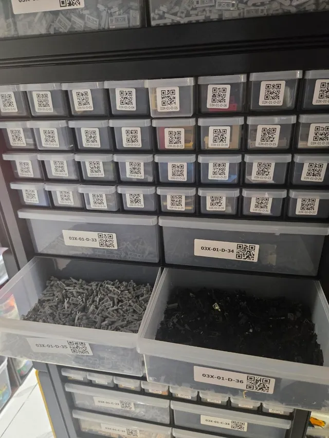 Two large drawers of dark gray and black LEGO parts pulled out of a black 36-drawer cabinet with QR labels