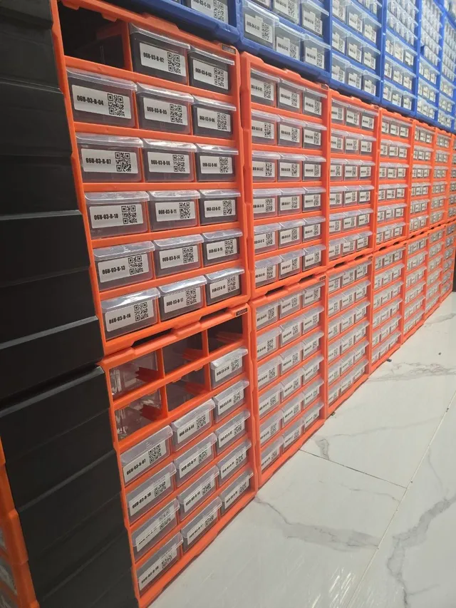 Aisle of orange VEVOR cabinets stacked into walls of QR-labeled drawers, blue cabinets on top, tile floor