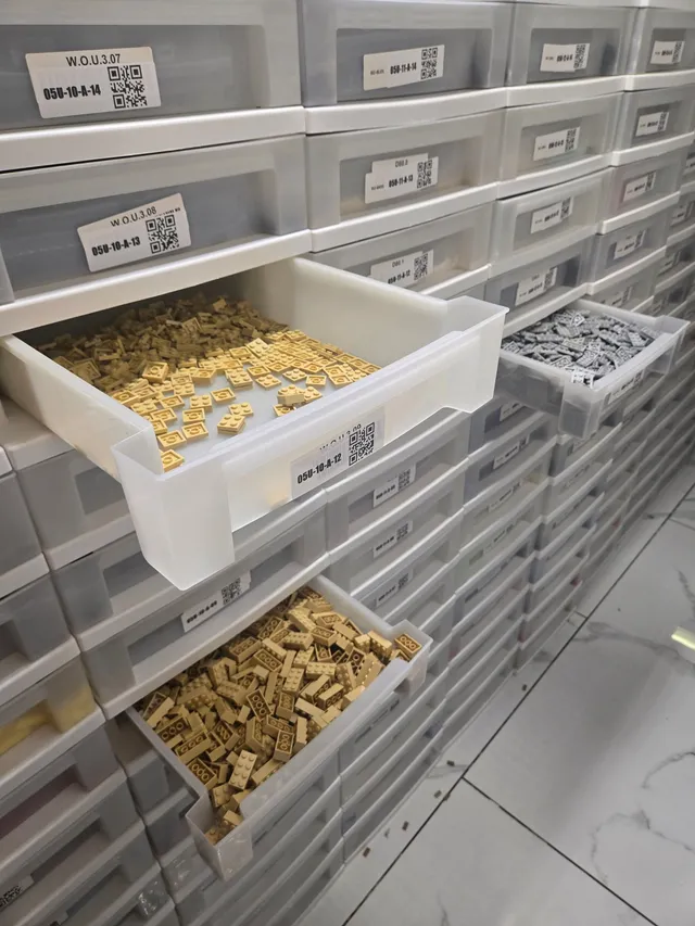 Three open drawers holding tan LEGO plates, tan bricks and gray plates in a wall of white QR-labeled drawers