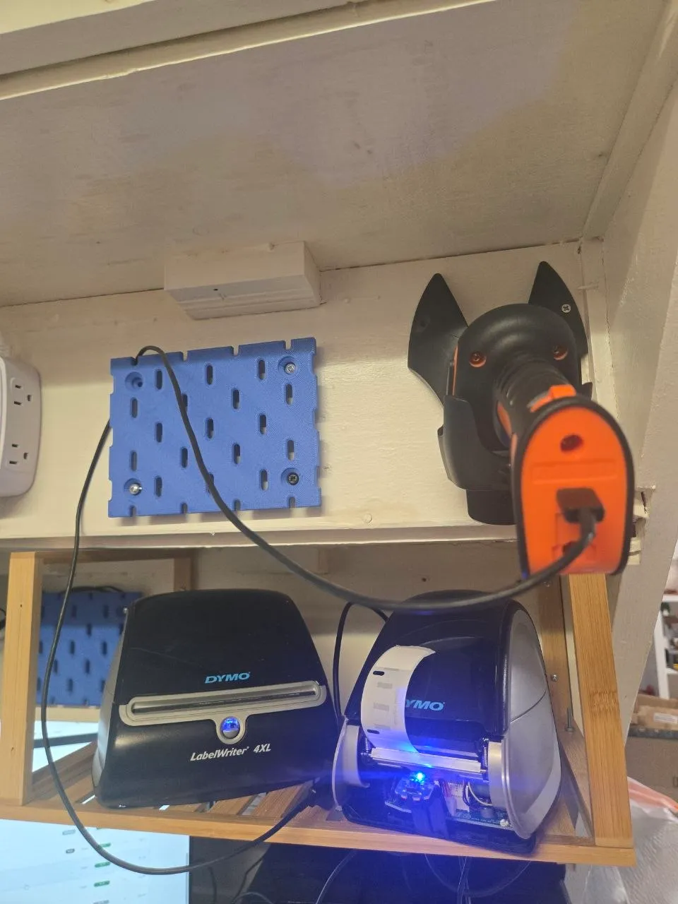 Black and orange Tera barcode scanner in a wall-mounted holder, USB cable plugged in, above two DYMO printers