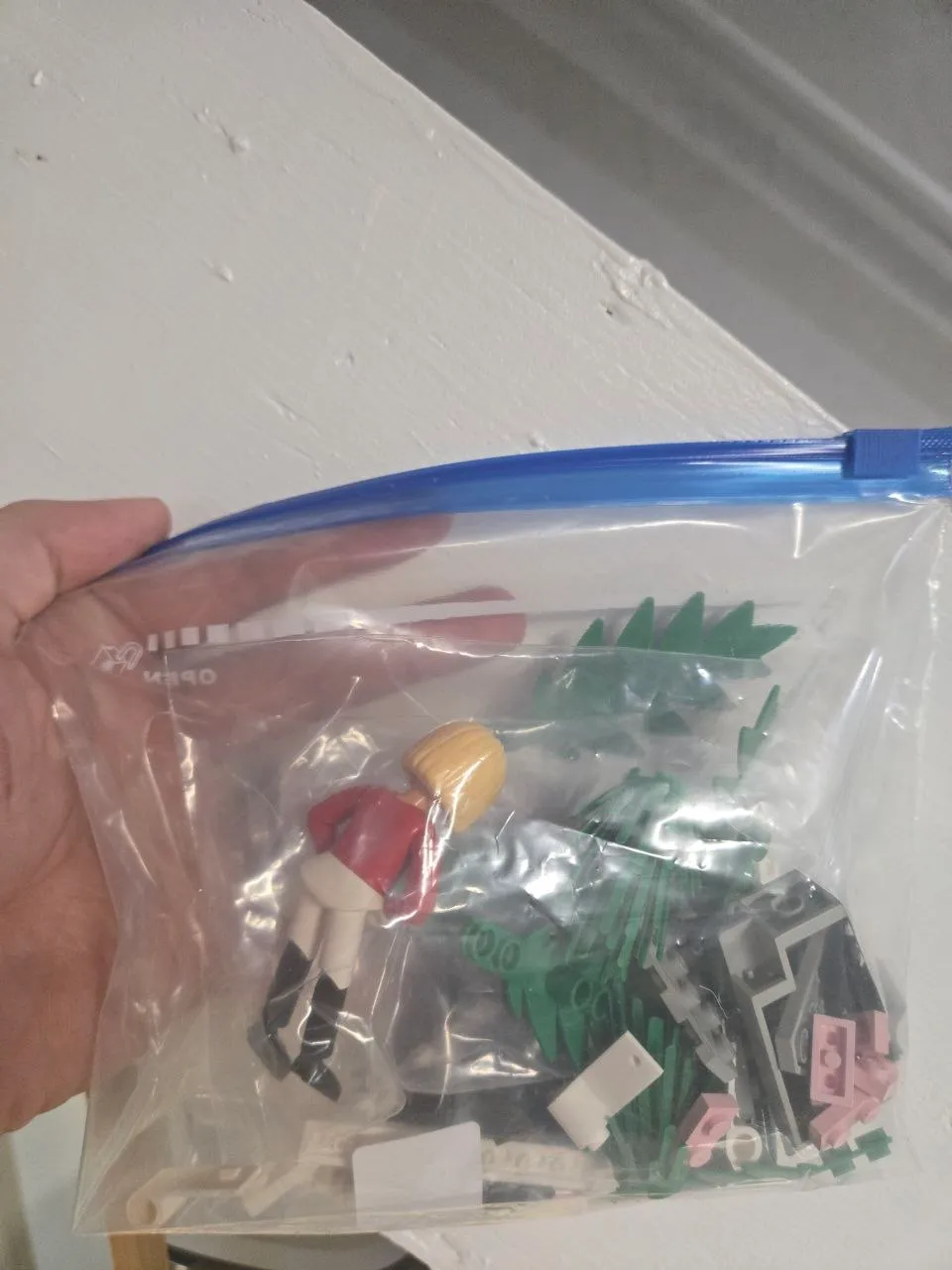 Hand holding a blue-slider bag with a LEGO doll in a red riding jacket, green plant pieces and pink bricks