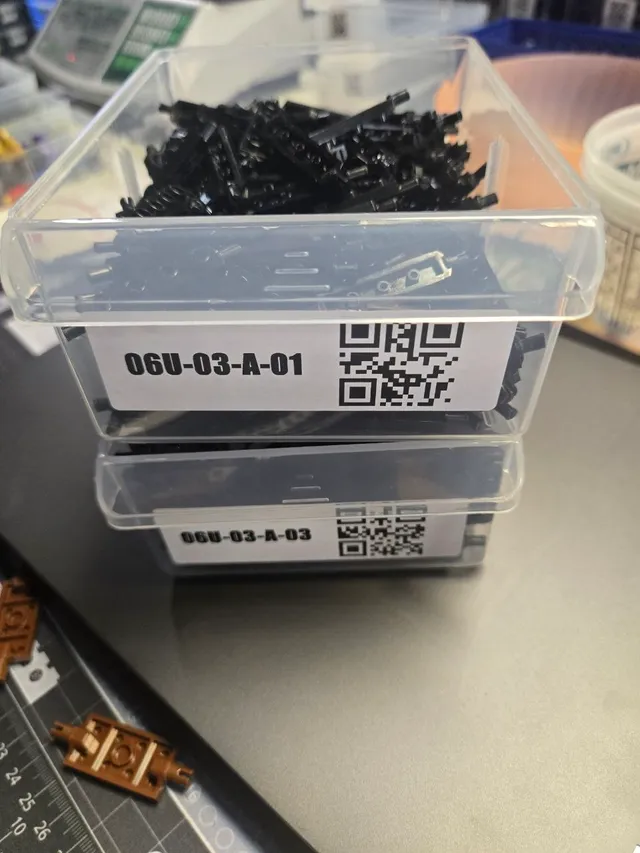 Two clear drawers taken out and stacked on a desk, the top one full of black LEGO parts, each with a QR label